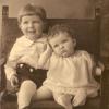 Two children sit on a chair facing the camera. The boy on the left is bigger and older than the girl on the right. The boy is smiling; the young girl rests her head on her hand.